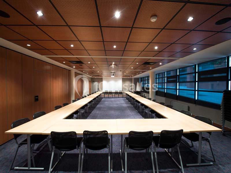Salle de réunion spacieuse avec tables disposées en U, chaises noires et éclairage au plafond.