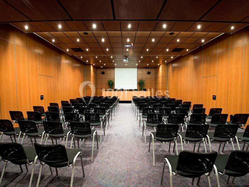 Salle de conférence vide avec rangées de chaises noires, un écran blanc et des murs en bois clair.