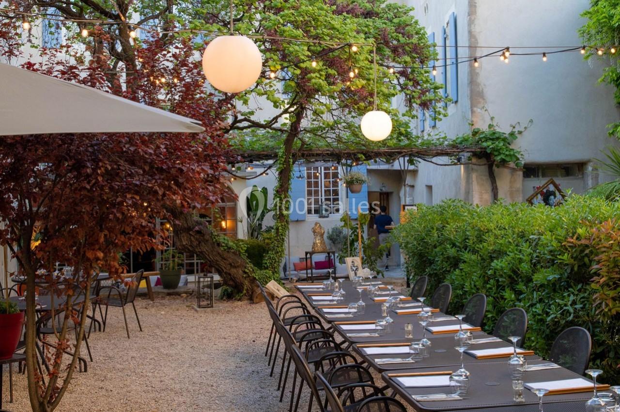 Terrasse extérieure avec tables dressées, éclairée par des guirlandes lumineuses et entourée de végétation.