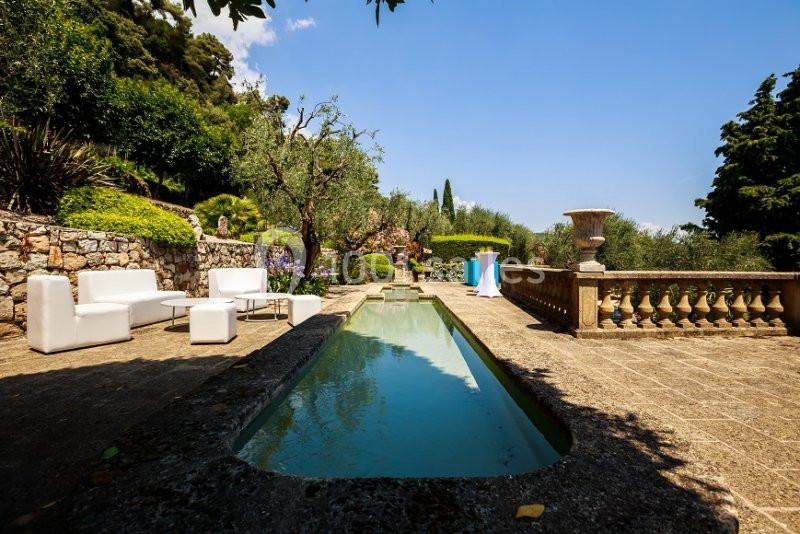 Terrasse en pierre avec piscine étroite, mobilier blanc, végétation méditerranéenne et ciel dégagé en arrière-plan.