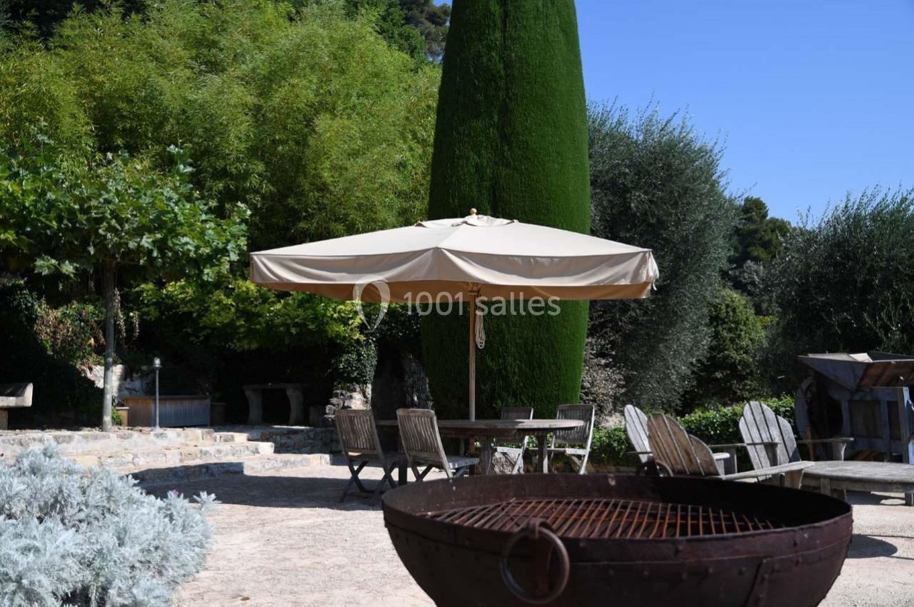 Espace extérieur avec parasol beige, chaises en bois, végétation dense et un brasero au premier plan.