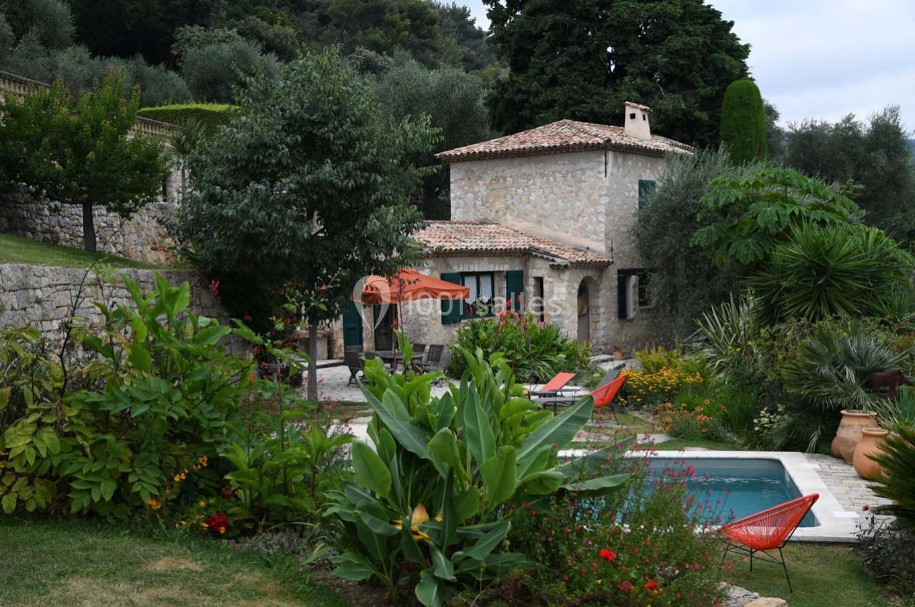 Maison en pierre entourée de végétation luxuriante avec une piscine et des chaises colorées dans un jardin.