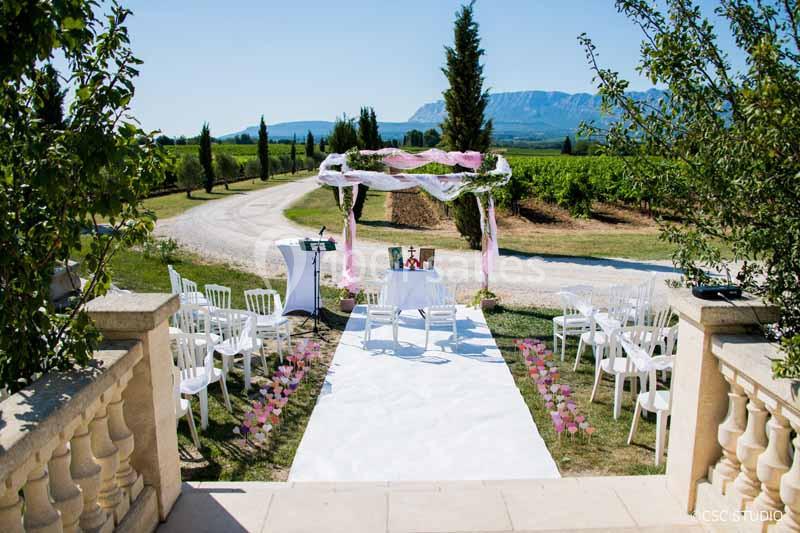 Cérémonie de mariage en extérieur avec arche décorée, chaises blanches et allée bordée de pétales, dans un cadre champêtre.