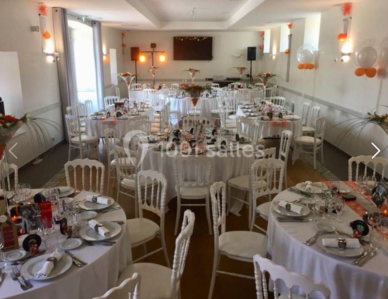 Salle de réception décorée avec des tables rondes dressées, chaises blanches et ornements orange et blancs.