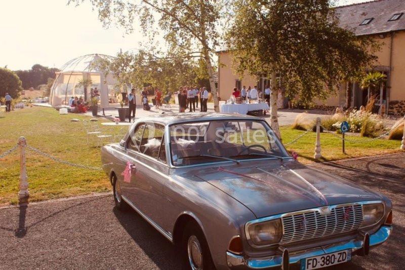 Voiture ancienne grise garée devant une salle de réception avec des invités et une structure transparente en arrière-plan.