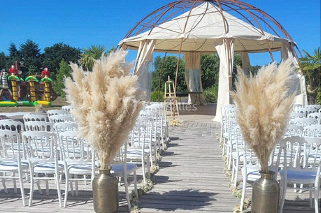 Allée centrale bordée de chaises blanches et de bouquets de pampas menant à une arche en toile sous un ciel bleu.
