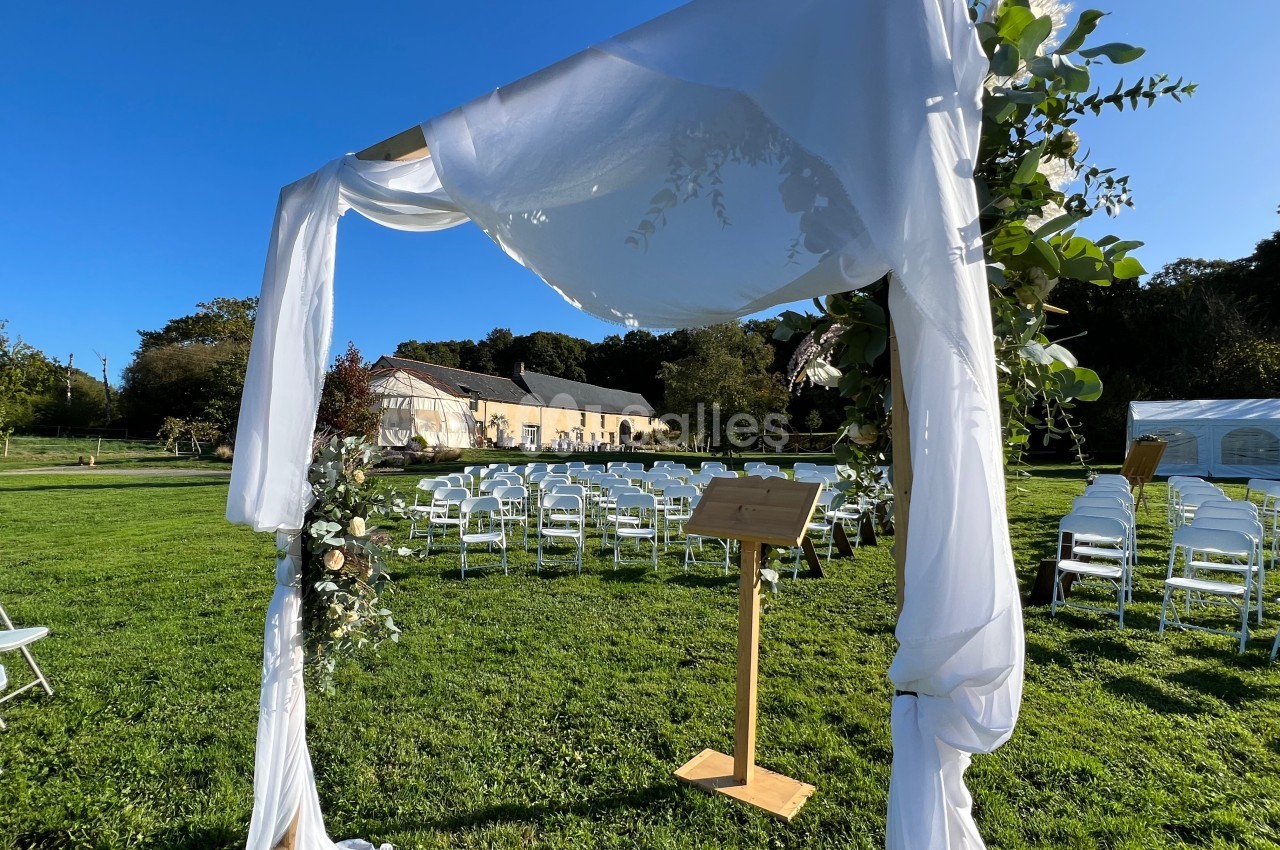 Arche de cérémonie décorée de tissu blanc et de feuillage, installée dans un jardin avec des chaises alignées en arrière…
