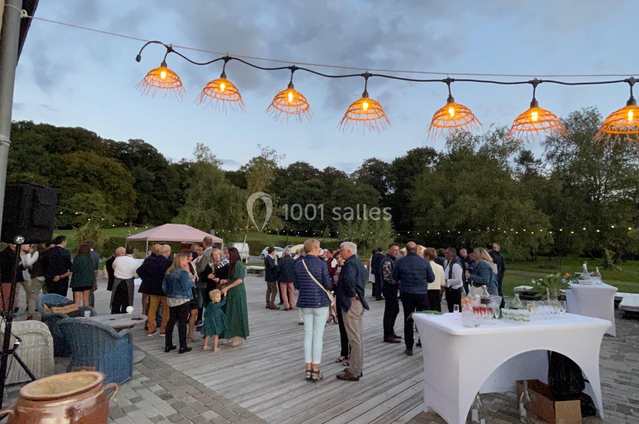 Groupe de personnes réunies sur une terrasse extérieure en soirée, sous des lampes suspendues, avec un jardin en arrière…