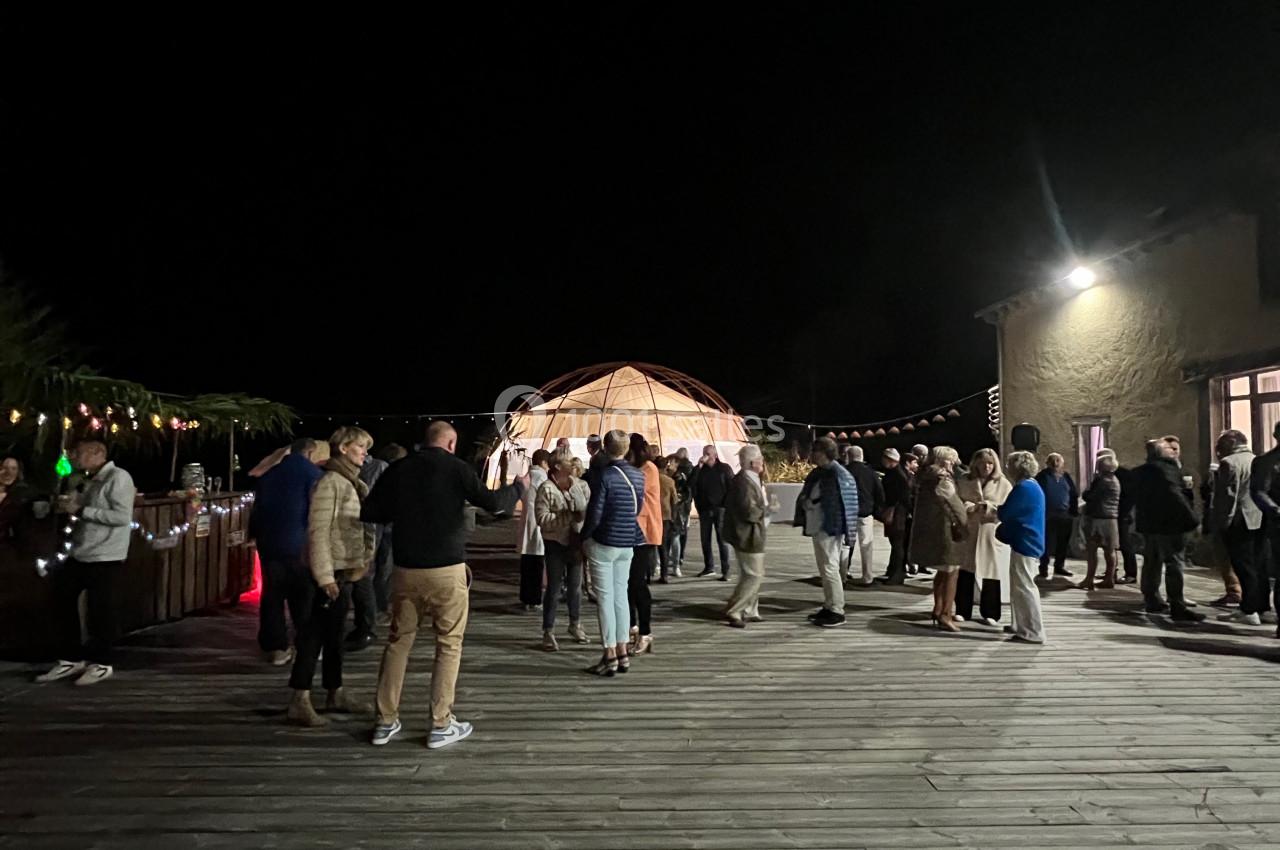 Groupe de personnes rassemblées sur une terrasse en bois la nuit, près d'une structure lumineuse et d'un bâtiment.