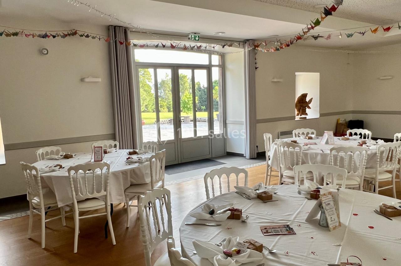Salle de réception lumineuse avec tables rondes dressées, décorée de guirlandes et ouverte sur un espace extérieur verdoyant.