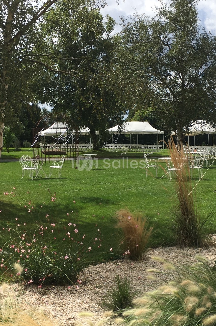 Chaises et tables blanches disposées sur une pelouse entourée d'arbres, avec des tentes blanches en arrière-plan.