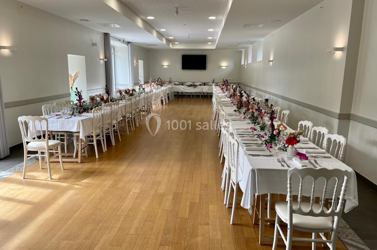 Salle lumineuse avec tables alignées et décorées de fleurs pour un événement, chaises blanches et parquet au sol.