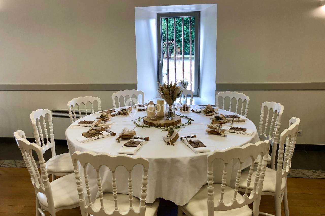 Table ronde décorée pour un événement, avec nappes blanches, chaises assorties et centre de table champêtre.