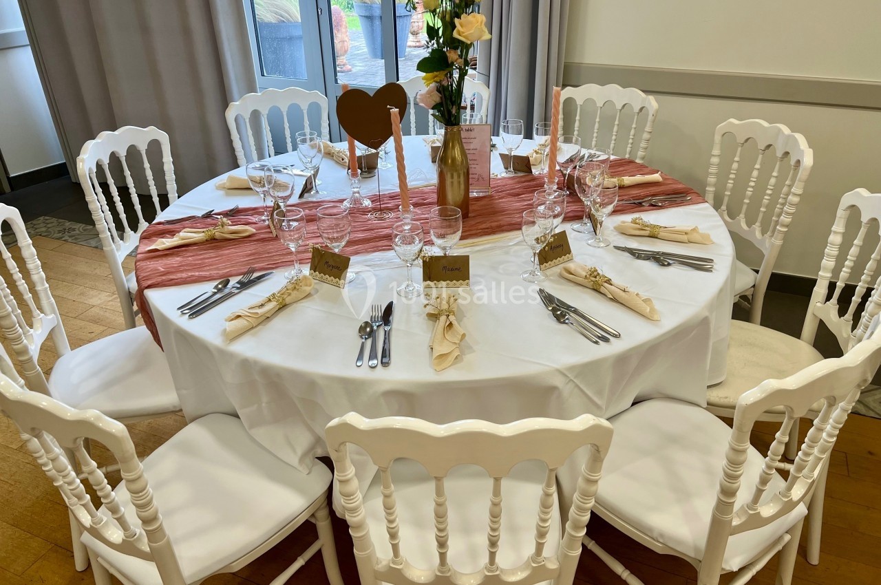 Table ronde décorée pour un événement, avec nappes blanches, chemins de table roses, vaisselle et chaises blanches.
