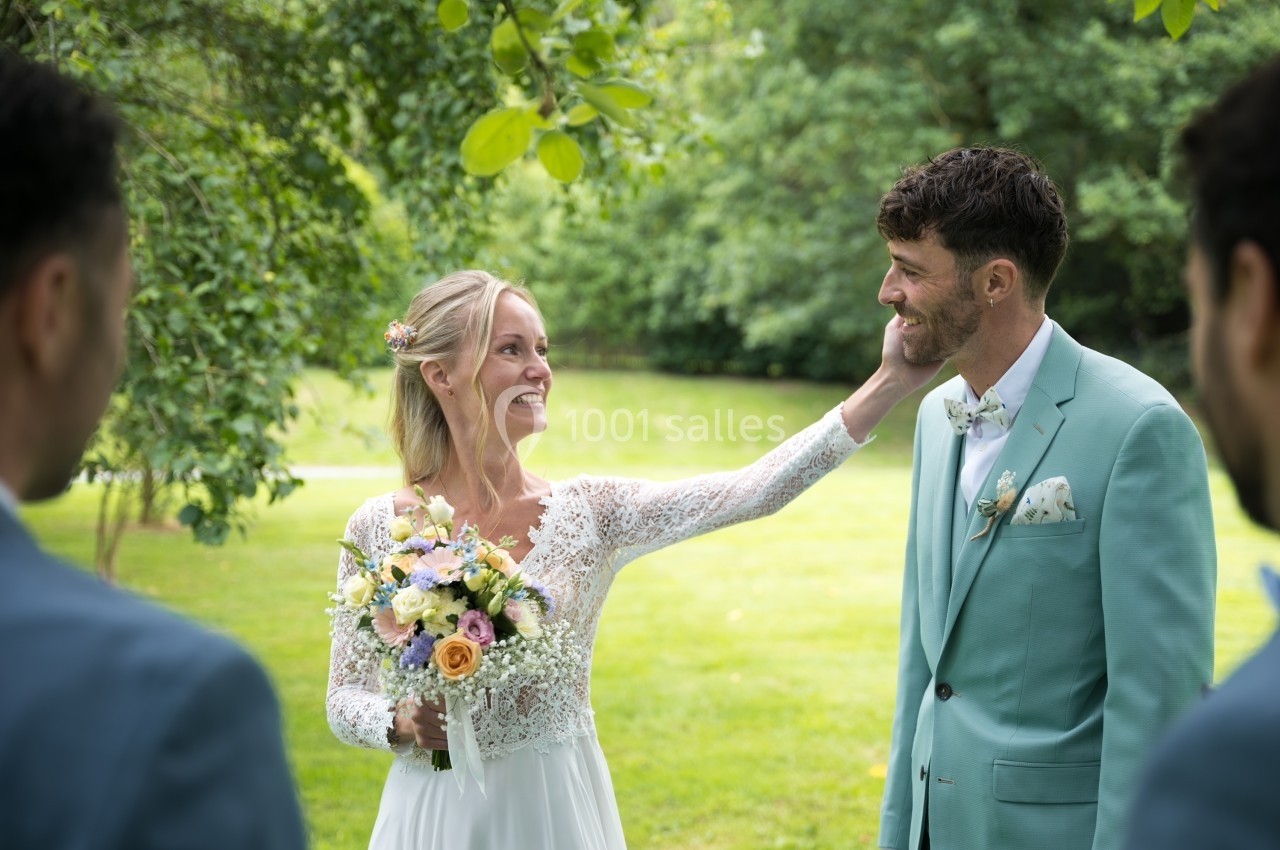 Une mariée souriante touche le visage du marié dans un jardin verdoyant, entourés de deux invités en costume.