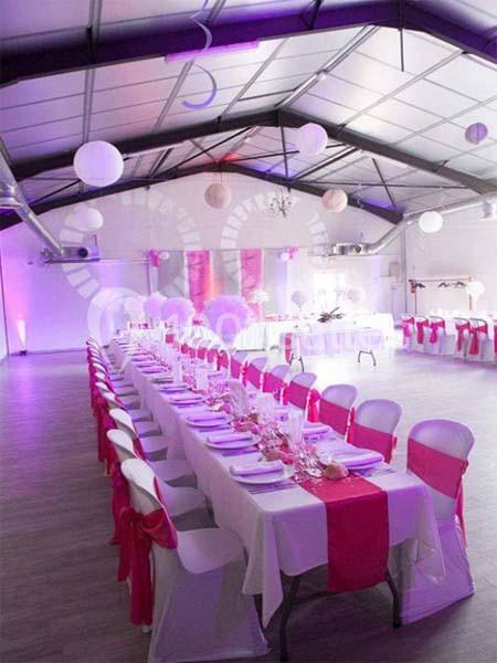 Salle décorée pour un événement avec longues tables dressées, nappes blanches, chemins de table roses et chaises assorties.