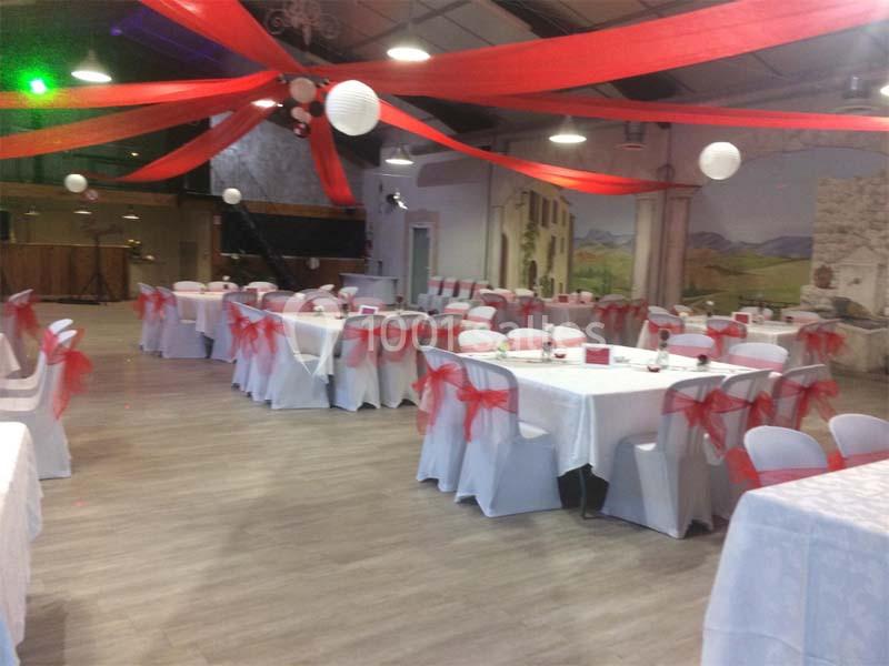 Salle de réception décorée avec des nappes blanches, des chaises ornées de nœuds rouges et des rubans suspendus.