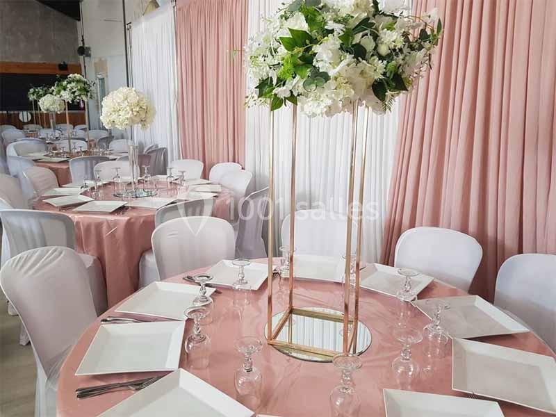 Salle de réception décorée avec des tables rondes, nappes roses, chaises blanches et centres de table fleuris.