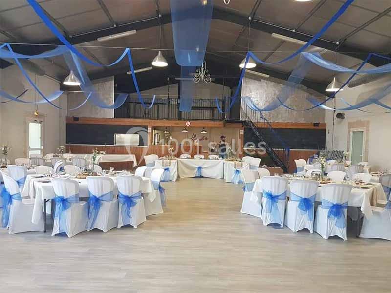 Salle de réception décorée de rubans bleus, avec des tables rondes dressées et des chaises blanches ornées de nœuds bleus.