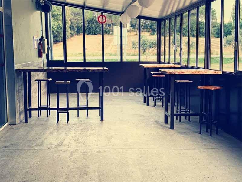 Salle lumineuse avec tables hautes en bois et tabourets, entourée de baies vitrées donnant sur un paysage arboré.
