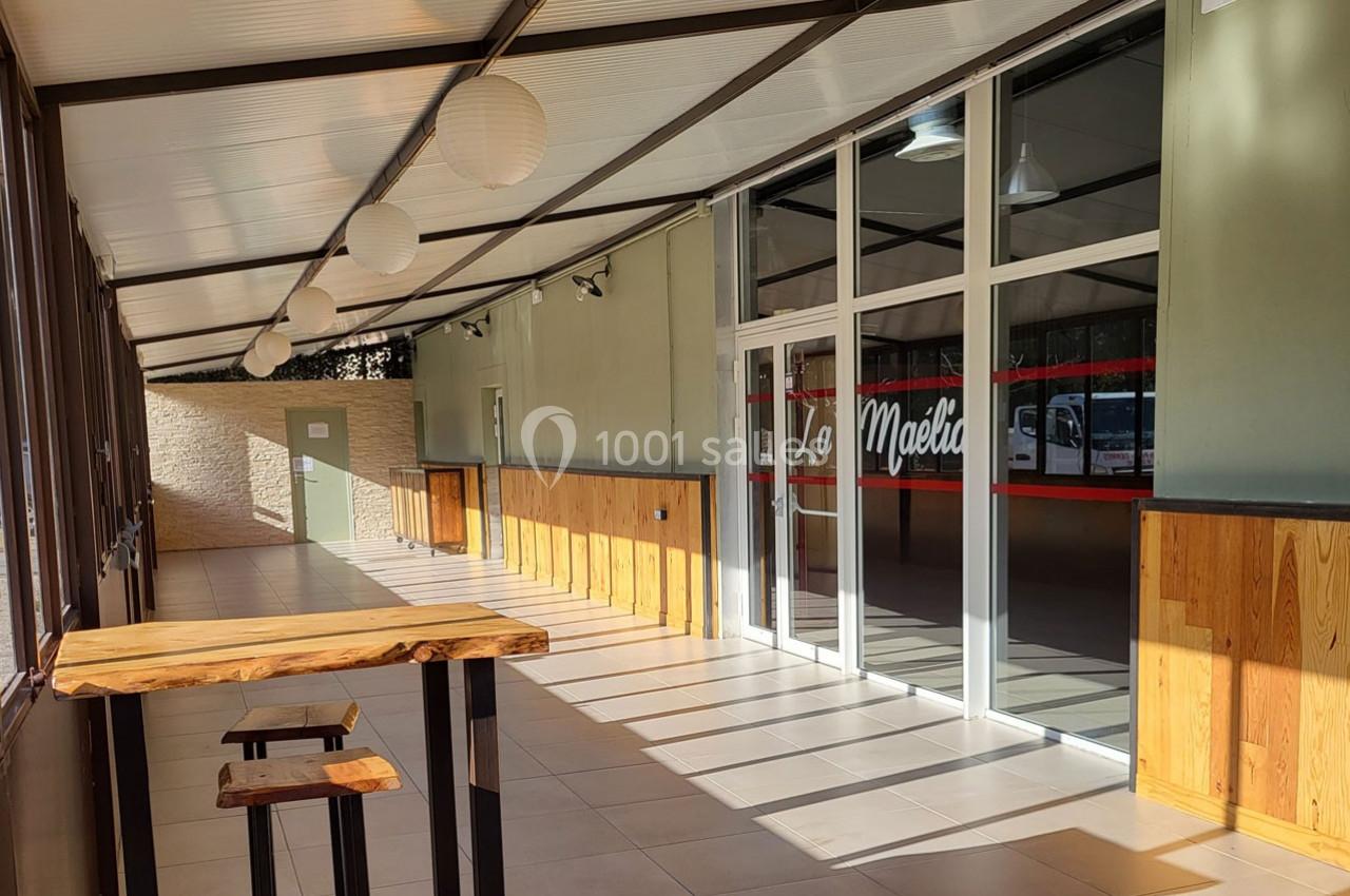 Terrasse couverte avec tables en bois, murs verts et entrée vitrée d'un bâtiment éclairé par la lumière naturelle.