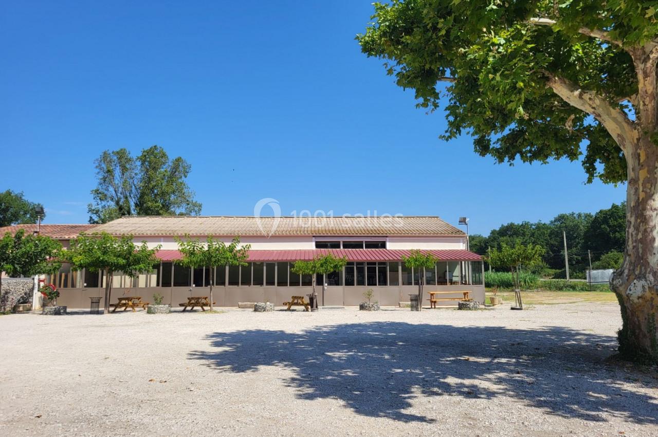 Bâtiment avec façade vitrée, terrasse ombragée par des arbres et tables en bois, situé sur une grande esplanade.