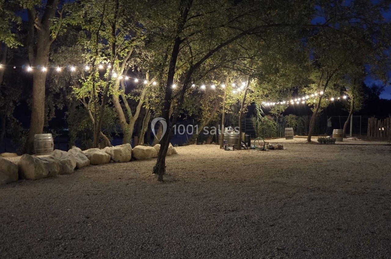 Cour extérieure en soirée, éclairée par des guirlandes lumineuses suspendues entre des arbres et entourée de pierres.