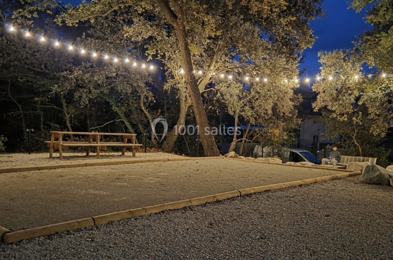 Terrain de pétanque en gravier éclairé par des guirlandes lumineuses, entouré d'arbres et de bancs en bois.