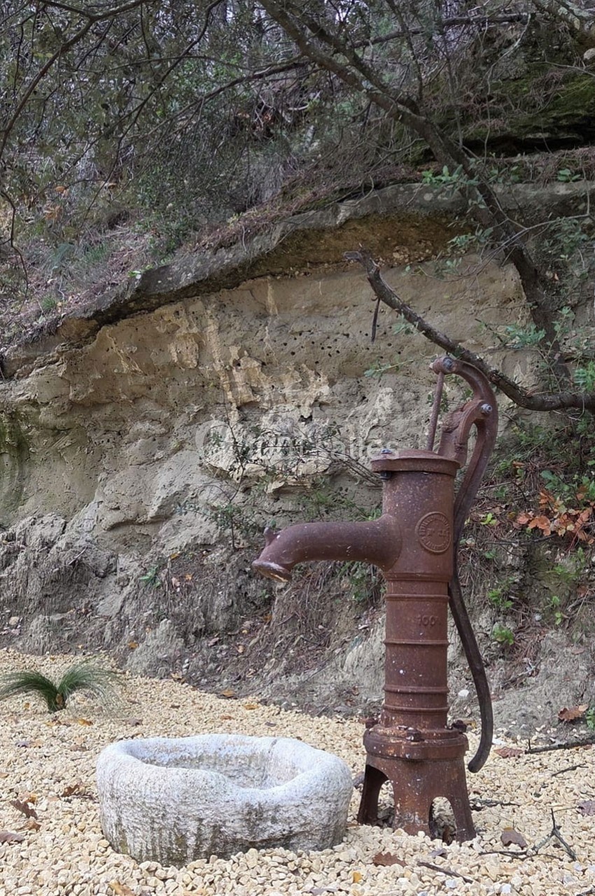 Pompe à eau en métal rouillé près d'un bassin en pierre, entourée de gravier et de végétation naturelle.