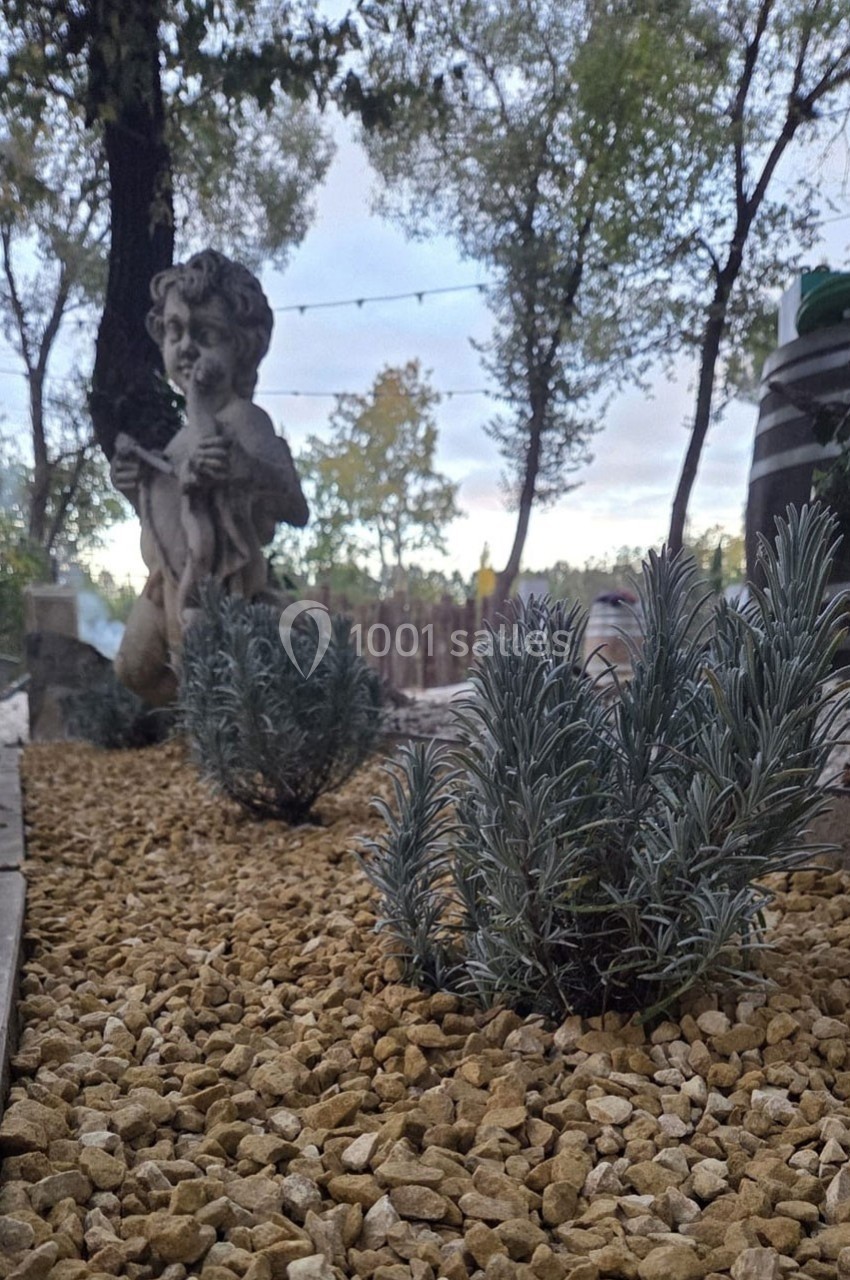 Statue décorative dans un jardin avec gravier, plantes et arbres en arrière-plan sous un ciel nuageux.