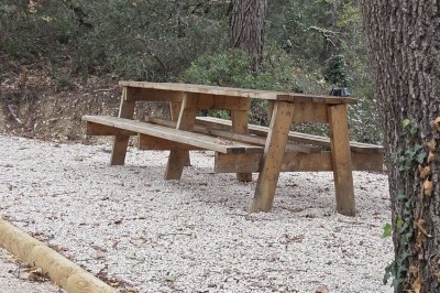Table de pique-nique en bois sur un sol de gravier, entourée d'arbres dans un cadre naturel.