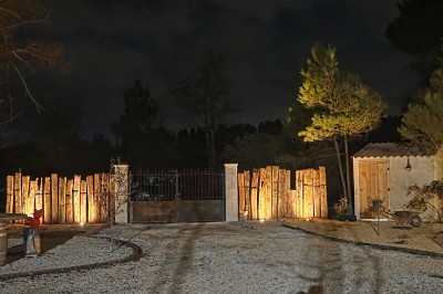 Clôture métallique devant un mur de pierre éclairé, entouré de végétation dans une ambiance nocturne.