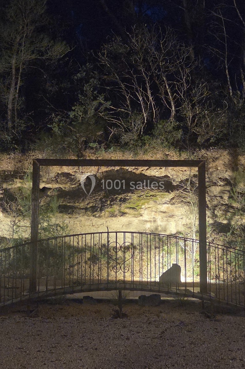 Clôture métallique devant un mur de pierre éclairé, entouré de végétation dans une ambiance nocturne.
