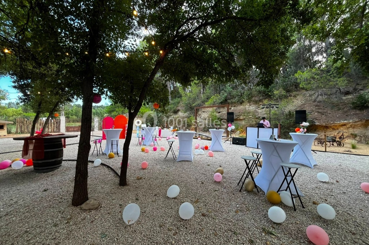 Location salle Loriol-du-Comtat (Vaucluse) - La Maelia #25 Espace extérieur décoré pour un événement, avec tables hautes, ballons colorés et guirlandes lumineuses sous les arbres.