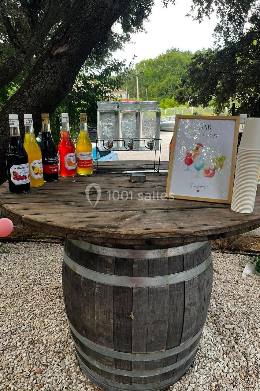 Location salle Loriol-du-Comtat (Vaucluse) - La Maelia #26 Table en tonneau avec sirops colorés, distributeurs d'eau, gobelets et affiche, installée en extérieur sous des arbres.
