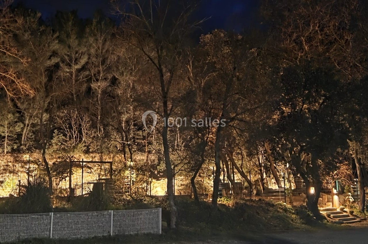 Location salle Loriol-du-Comtat (Vaucluse) - La Maelia #32 Vue nocturne d'un espace boisé éclairé par des lumières artificielles, avec des arbres et des structures en arrière-plan.