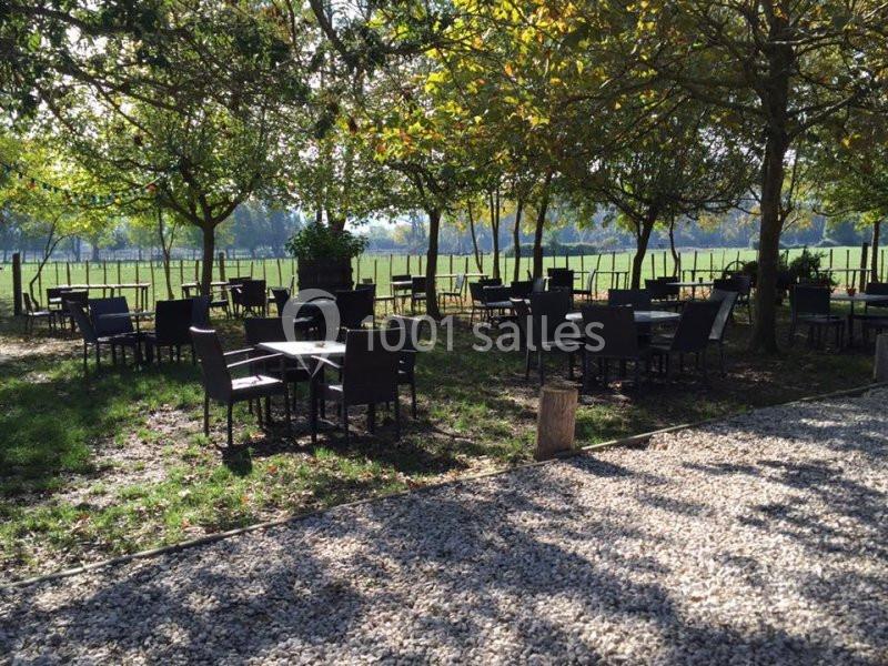 Terrasse extérieure avec tables et chaises sous des arbres, donnant sur un champ verdoyant par temps ensoleillé.
