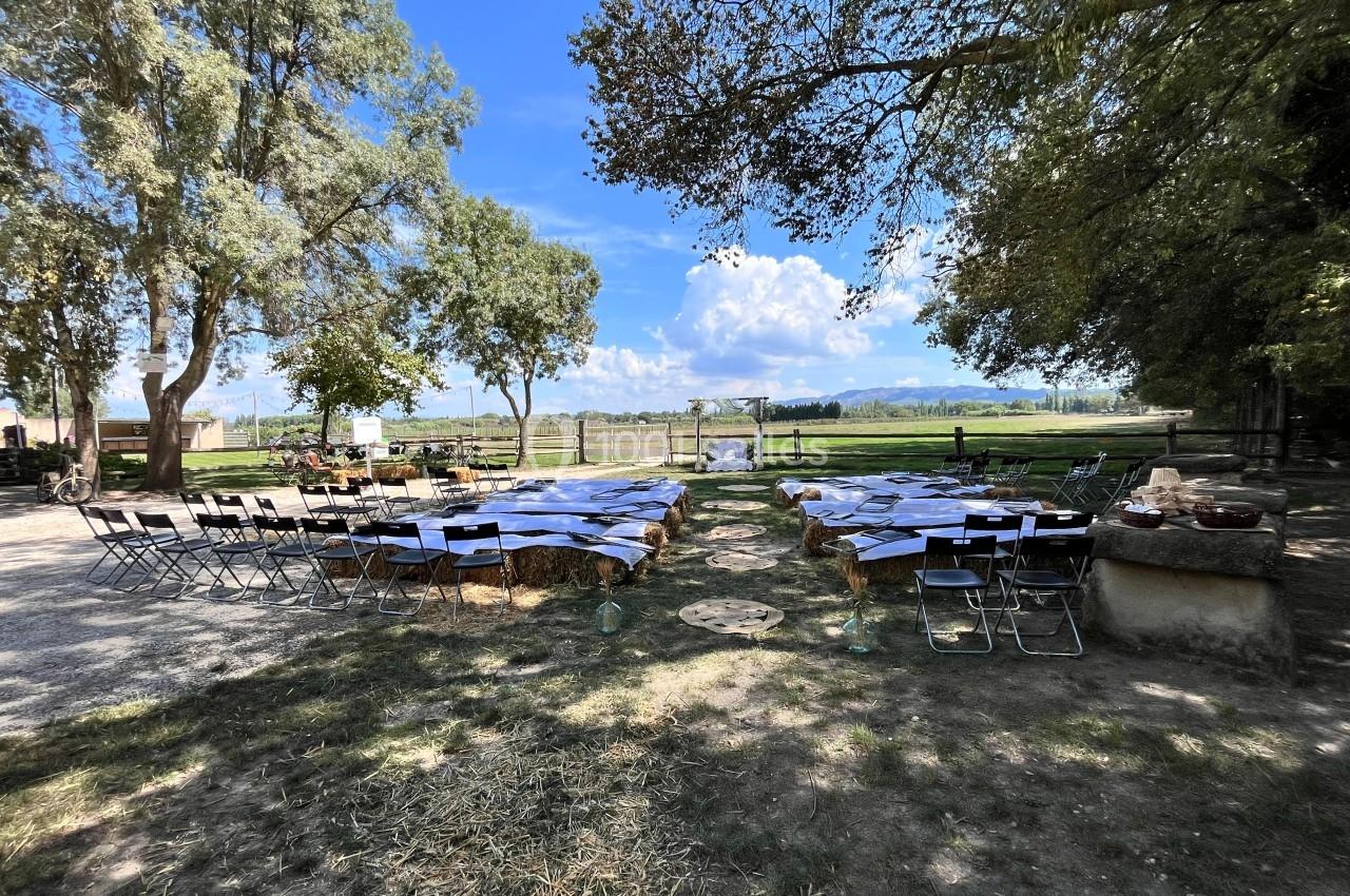Chaises disposées en extérieur devant une arche décorée, entourées d'arbres et de champs sous un ciel bleu.