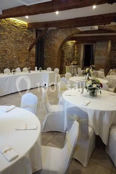 Salle en pierre avec poutres apparentes, tables rondes et rectangulaires décorées pour un événement.