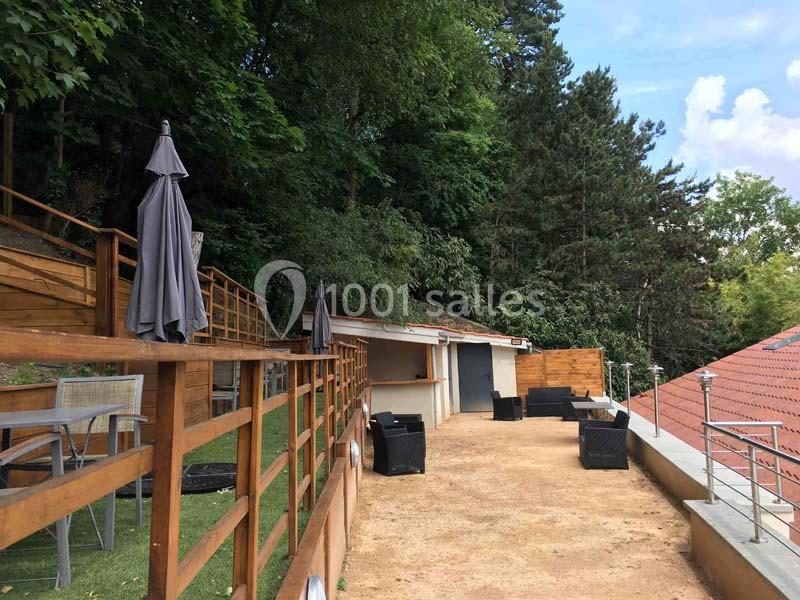 Terrasse extérieure avec tables, chaises et parasols, entourée de verdure et surplombant un toit en tuiles.