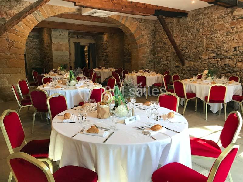 Salle en pierre avec des tables rondes dressées pour un repas, chaises rouges et décoration florale.