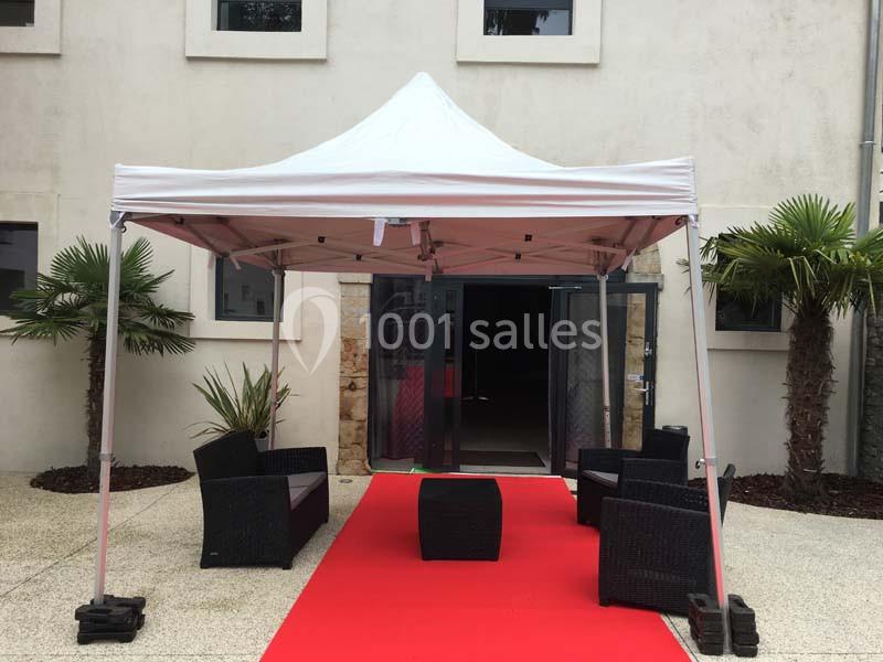 Tente blanche avec mobilier de jardin noir sur tapis rouge, installée devant une entrée vitrée d'un bâtiment beige.
