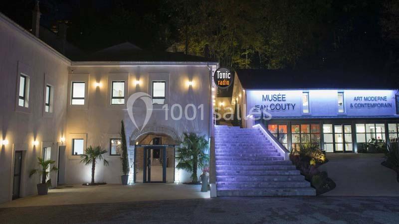 Façade éclairée du musée Jean Couty avec escalier central et enseigne lumineuse, entourée de végétation.