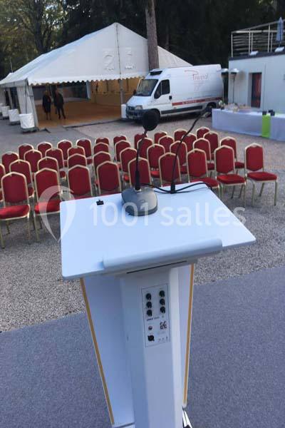 Location salle Lyon 9 (Rhône) - Hub de l'Ile Barbe #22 Un pupitre équipé de micros devant des rangées de chaises rouges disposées en extérieur, près d'une tente blanche.