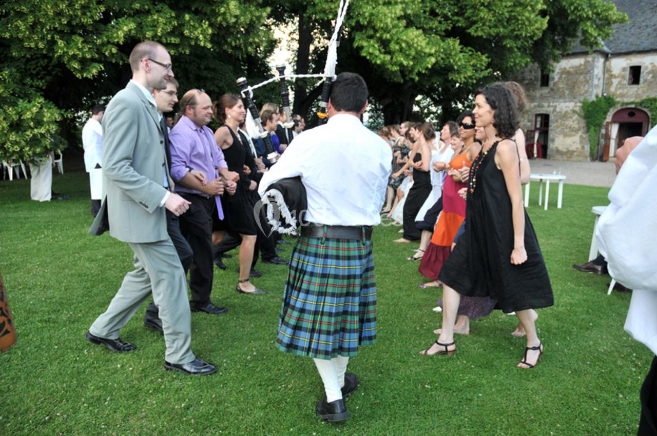 Personnes dansant en cercle sur une pelouse, guidées par un joueur de cornemuse en kilt, lors d'un événement en extérieur.