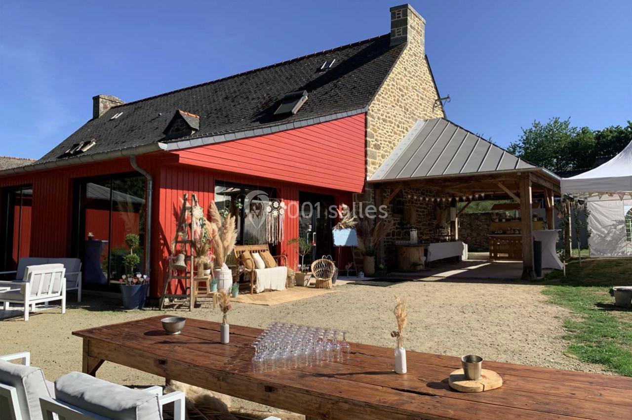Maison en pierre avec extension rouge, terrasse aménagée, grande table en bois et espace couvert dans un jardin ensoleillé.