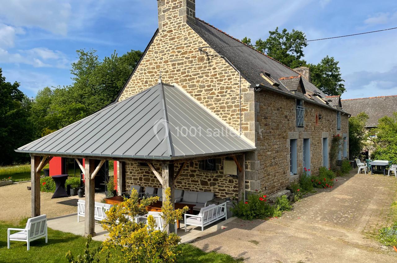Maison en pierre avec toit en ardoise, terrasse couverte, mobilier de jardin et environnement verdoyant.