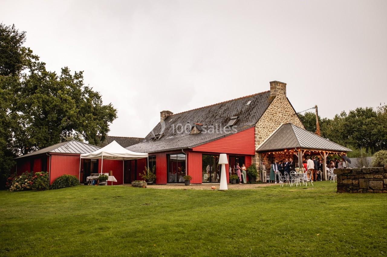 Maison en pierre et bois rouge avec toit en ardoise, entourée de verdure, des personnes rassemblées sous un auvent.