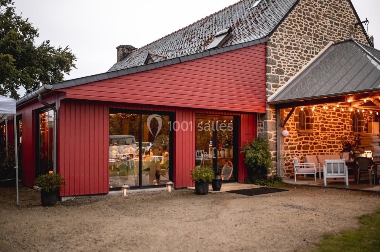 Façade d'une maison en pierre avec une extension en bois rouge, éclairée par des lumières chaleureuses en soirée.