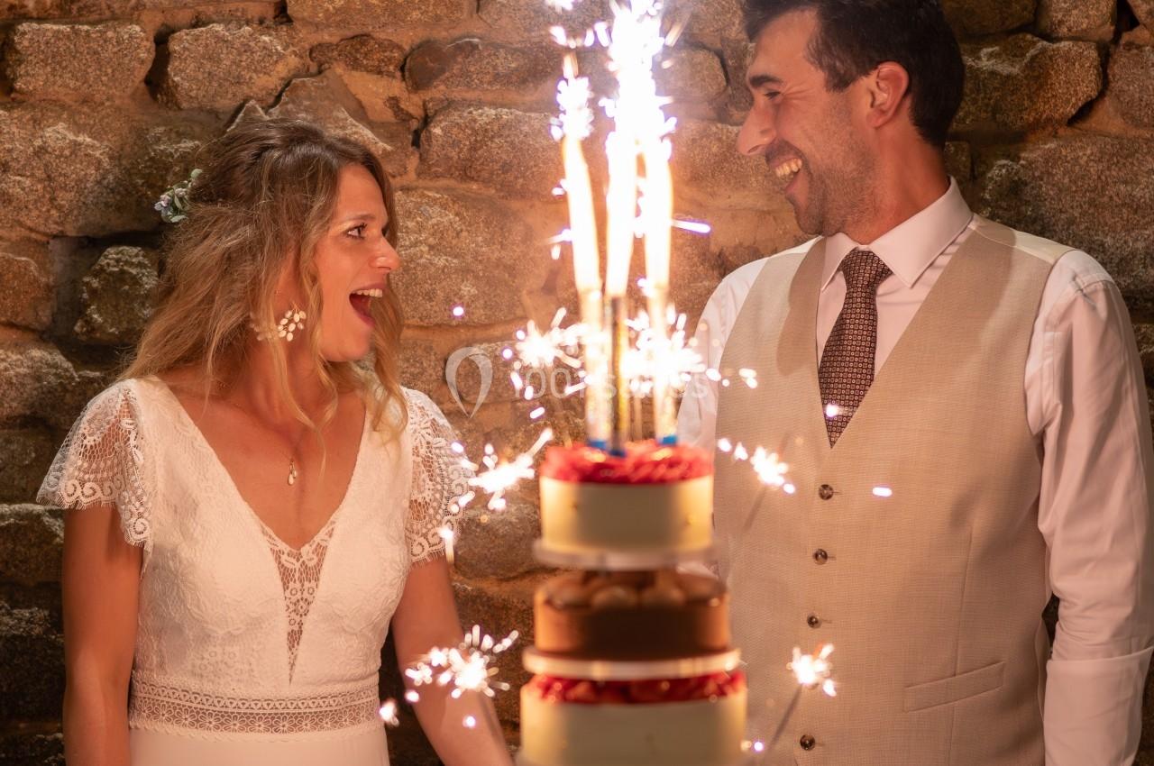 Un couple souriant devant une pièce montée décorée de cierges étincelants, sur fond de mur en pierre.